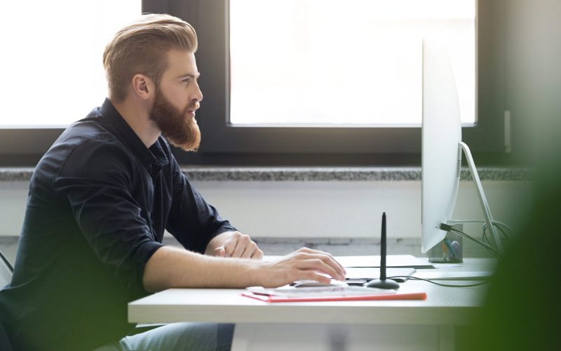 Side view of a young bearded man sitting at his desk and working on a computer